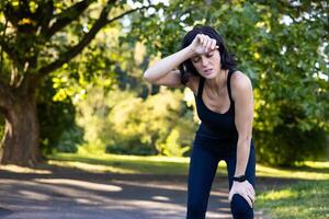 Young active woman in sports black clothes standing in park and bent over from fatigue after training, running and marathon, wiping sweat with hand from forehead and resting. photo