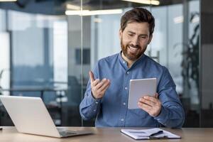 A young man sits in the office at a desk with a laptop and smilingly talks on a call on a tablet. photo