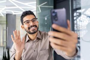 Joyful professional man engaging in a call using his smartphone in a contemporary office setting. He waves happily, radiating positivity and confidence. photo