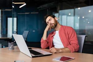 Tired businessman in casual red shirt sleeps at workplace, man on desk fell asleep during working hours inside office with laptop. photo