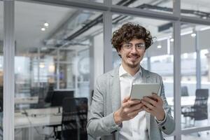 Professional young adult with curly hair, smart business attire, engaged with tablet inside bright office setting. photo