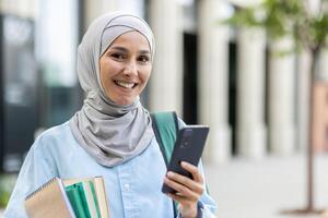 sonriente joven árabe mujer en hijab participación cuadernos y un teléfono inteligente fuera de un oficina, simbolizando empoderamiento y educación. foto
