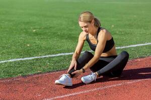 atlético mujer en ropa de deporte participación su tobillo en dolor en un brillante soleado día a el corriendo pista, representando Deportes lesión. foto