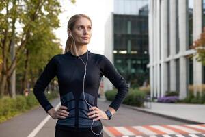 A fitness-focused woman pauses during her run in the city, displaying determination and confidence. Wearing sportswear and earphones, she embodies an active lifestyle against an urban backdrop. photo
