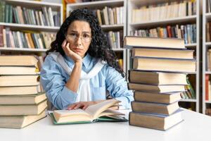 preocupado Hispano hembra estudiante estudiando difícil en un biblioteca. ella es rodeado por grande pilas de libros y mira abrumado. foto