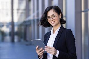 de cerca retrato de un joven negocio mujer en pie fuera de un oficina edificio, participación un tableta y sonriente a el cámara. foto