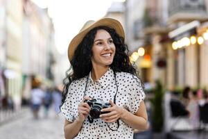 Young beautiful hispanic woman with curly hair walking in the evening city with a camera, female tourist on a trip exploring historical landmarks in the city. photo