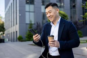 Cheerful Asian male executive enjoying a coffee break outside modern office buildings, using his phone with earphones. photo