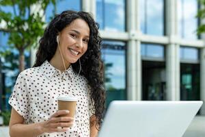 sonriente joven mujer con auriculares y café tomando un descanso desde trabajo en su ordenador portátil fuera de un moderno oficina edificio. foto