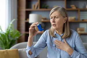 An elderly woman experiences an asthma attack and uses a blue inhaler. She looks concerned while sitting in a modern living room. photo