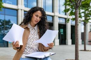 Worried young professional woman looking at documents with a puzzled expression outside a modern office building. photo