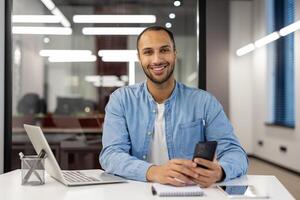 retrato de un sonriente joven Hispano musulmán hombre sentado en un moderno oficina a un escritorio, participación un móvil teléfono y mirando a el cámara. foto