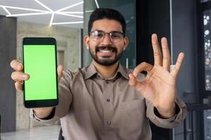 de cerca retrato de un joven indio hombre en pie en el oficina en frente de el cámara, sonriente y demostración un verde Bosquejo móvil teléfono pantalla y dedo gesto OK. foto