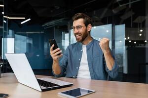 exitoso maduro empresario jefe trabajando dentro moderno oficina edificio a escritorio, utilizando computadora portátil, hombre con barba y lentes participación teléfono inteligente, tiene bueno Noticias acerca de victoria triunfo celebrando. foto