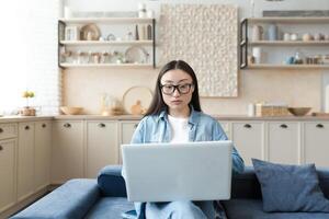 Young asian businesswoman working remotely from home sitting on sofa serious and focused using laptop for online freelance work photo