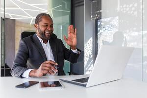 Joyful successful african american man inside office at workplace talking remotely on call, man smiling and looking at laptop screen, waving hand in greeting gesture. photo