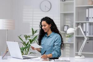 un alegre Hispano mujer gestiona papeleo en su moderno hogar oficina, exudando profesionalismo y positividad foto