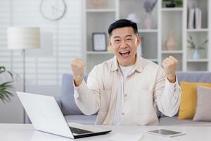 Portrait of successful Asian businessman mature man working remotely from home freelancer, living room sitting at table looking at camera and happy holding hands up celebrating victory and triump. photo