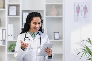 Doctor inside the medical office stands with a tablet computer in hands, a clinic worker in white medical robe will advises the patient remotely using the for call, joyfully and friendly. photo