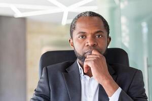 Mature focused african american man looking at camera, close up portrait of thoughtful concentrated boss at workplace inside office in business suit. photo