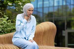 A young Muslim woman wearing a hijab looks uneasy as she sits on a wicker bench outside a modern building, clutching her throat and chest, indicating discomfort or illness. photo