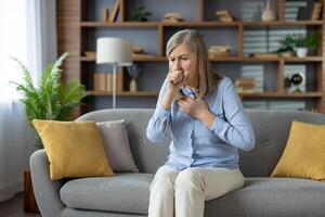 Senior stylish woman feeling sick and covering mouth with clenched fist for stopping cough while sitting on sofa with cushions. Unhealthy female feeling pain in chest and suffering from flu symptoms. photo