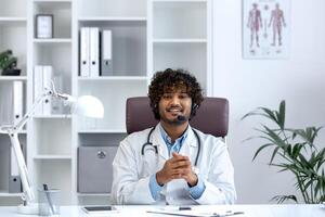 cámara web vista, joven médico con auriculares teléfono utilizando ordenador portátil para llamar, médico alegremente y simpático consultante pacientes, sonriente y mirando a cámara, trabajando dentro clínica médico habitación. foto