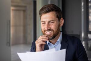 A smiling businessman in a suit examines papers, portraying confidence and professionalism in a well-lit office setting. photo