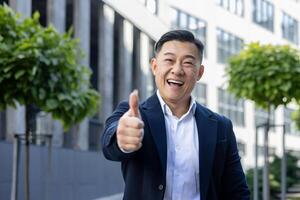 Cheerful Asian businessman in business attire giving a thumbs up, expressing approval and success, outdoor setting. photo