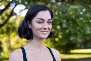 Close-up portrait of a young smiling woman in sporty black clothes standing outside in a park while exercising and jogging, and looking confidently at the camera. photo