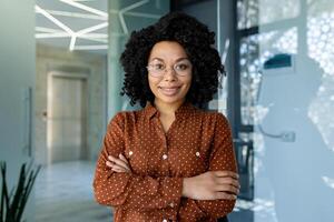 retrato de exitoso oficina obrero, africano americano mujer con Rizado pelo sonriente y mirando a cámara, mujer de negocios con cruzado brazos a lugar de trabajo contento con logro resultados dentro oficina foto