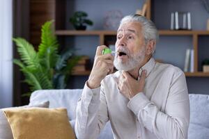 Elderly Caucasian man in a cozy living room using a throat spray. He looks concerned, suggesting discomfort possibly from a sore throat or cold symptoms. photo