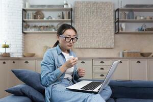 en línea consulta con psicólogo, un asiático mujer en lentes es solo a hogar llanto, sentado en el sofá vivo habitación utilizando un ordenador portátil para un llamada hablando apoyo cursos foto