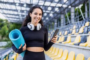 Portrait of happy female athlete, Hispanic woman standing near stadium outside smiling and looking at camera, holding phone and exercise mat, using app for co-training. photo