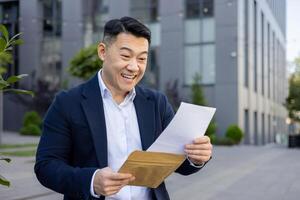 Happy Asian businessman reading a letter with excitement outside an office building, expressing success and joy. photo