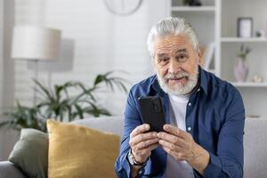 Cheerful elderly man with gray beard smiling as he looks at his smartphone, sitting comfortably on the couch at home. photo