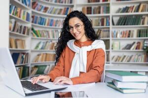 joven Hispano mujer estudiando en académico Universidad biblioteca, hembra estudiante sonriente y mirando a cámara mientras sentado a computadora portátil, mujer con Rizado cabello. foto