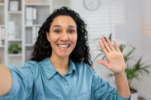 un alegre mujer es ondulación y sonriente a el cámara, representando un simpático saludo o diciendo adiós durante un en línea llamada desde hogar. foto