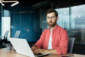 Serious and focused mature businessman in red shirt wearing glasses looking at camera, young man at work typing on laptop keyboard, programmer developer portrait at work. photo