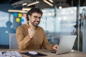 Cheerful businessman with glasses celebrating a success while working on his laptop in a modern office environment. photo