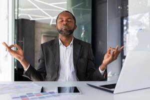 Mature experienced african american boss meditating inside office workplace sitting at desk, man with closed eyes sitting visualizing future wins and achievements, businessman investor with laptop. photo