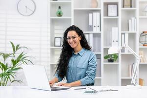 A joyful Latina businesswoman working remotely from a well-organized home office, engaging confidently with the camera. photo