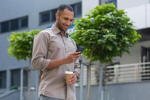 africano americano empresario sonriente y contento caminando fuera de el oficina edificio, participación un taza de caliente bebida durante un romper, y utilizando el teléfono, hojeada web paginas y mecanografía mensajes foto