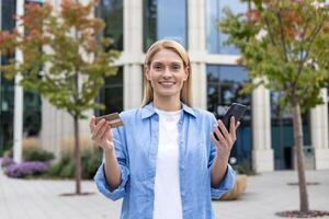 satisfecho caucásico mujer que lleva Teléfono móvil y el plastico crédito tarjeta mientras dejando financiero empresa edificio. rico sonriente dama recepción banco cuenta para especial clientes con prima sistema. foto