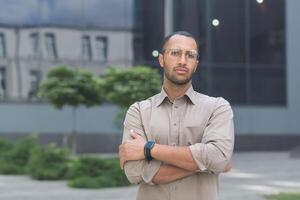 Serious and pensive young office worker with crossed arms looking at camera, businessman in shirt outside office building pensive photo