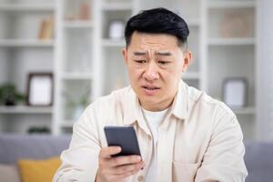 Upset Chinese man sitting in bright living room, holding phone in hands, sad because of bad news, smartphone not working, missed important call. photo