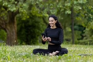 Smiling woman dressed in black activewear sitting on grass, engaging with her smartphone in a lush green park. photo