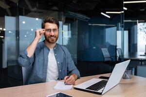 retrato de exitoso empresario detrás papel trabajar, hombre en camisa sonriente y mirando a cámara, financiero jefe dentro oficina utilizando ordenador portátil en trabajo a lugar de trabajo. foto