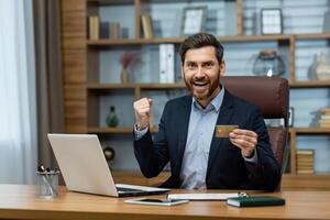 Joyful mature businessman holding a credit card and celebrating a financial triumph in a tastefully decorated home office. photo