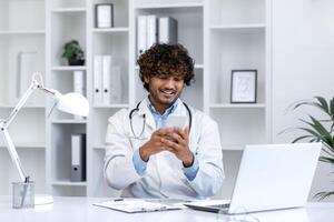 joven exitoso Hispano médico utilizando teléfono, hombre leyendo educativo material utilizando aplicación en teléfono inteligente, trabajando dentro clínica médico oficina, sonriente contentamente, sentado con ordenador portátil. foto